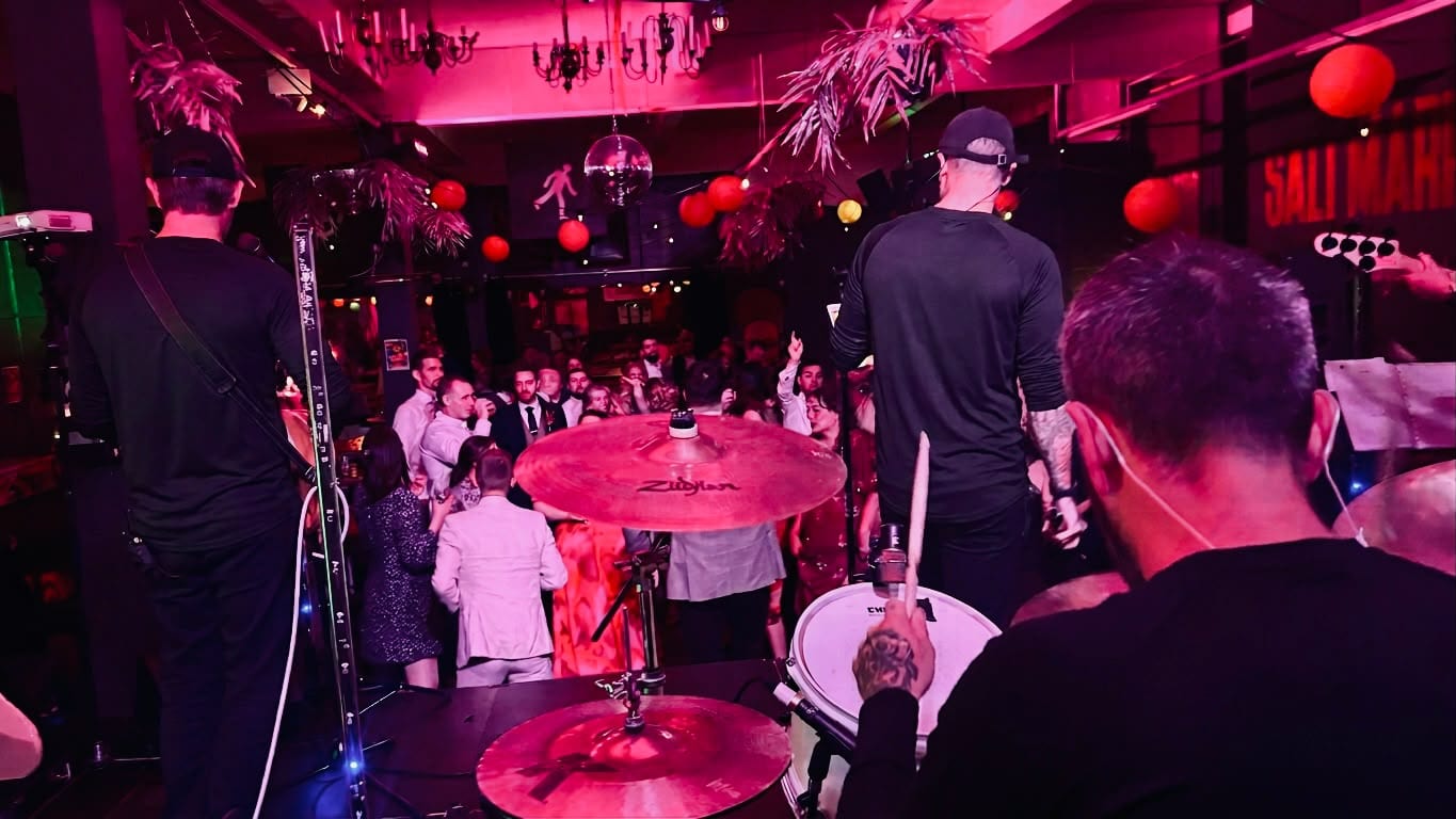 The view from behind the drummer out into a crowded room lit deep pink